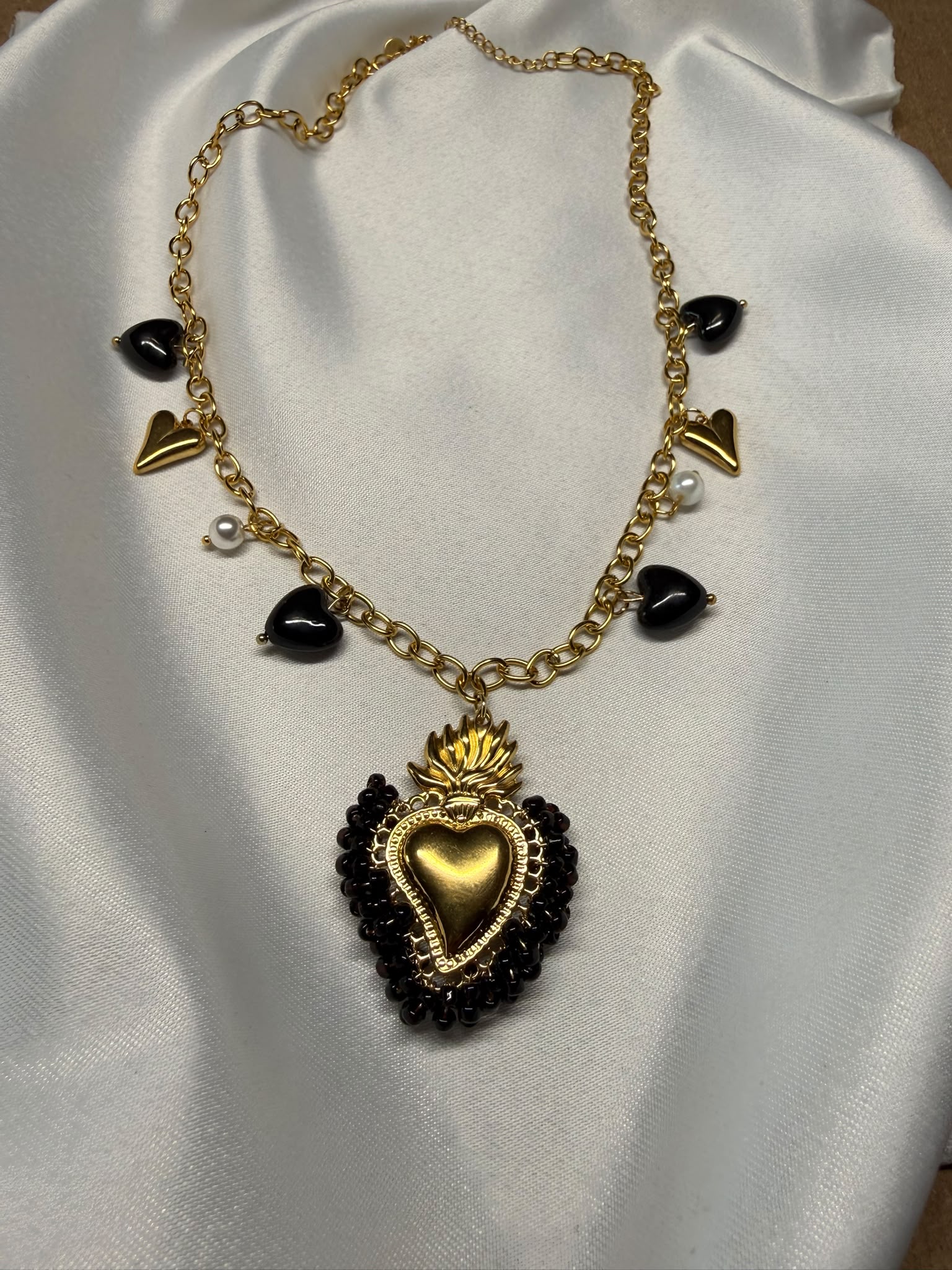 Collana con pendente Sacro Cuore dorato decorato con perline nere, su catena a maglie dorate con charm cuori neri, cuori dorati e perle bianche